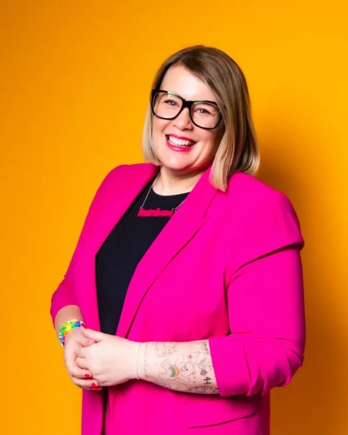 Rainbow Tomes smiling, in a bright pink suit against a yellow background.