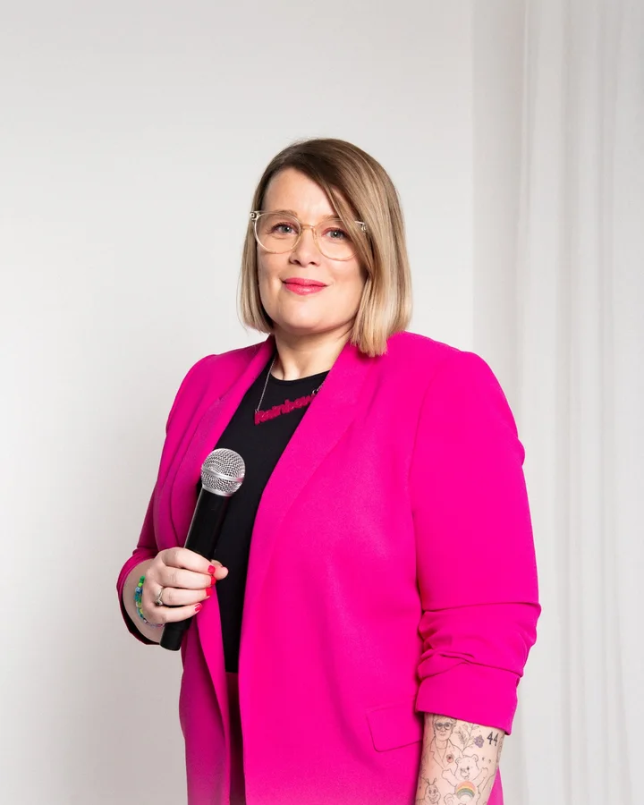 Rainbow in a pink jacket, smiling and holding a microphone.