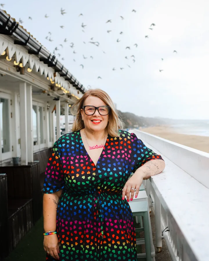 Rainbow smiling on a seafront terrace in a dress covered in rainbow hearts, with birds flying overhead.